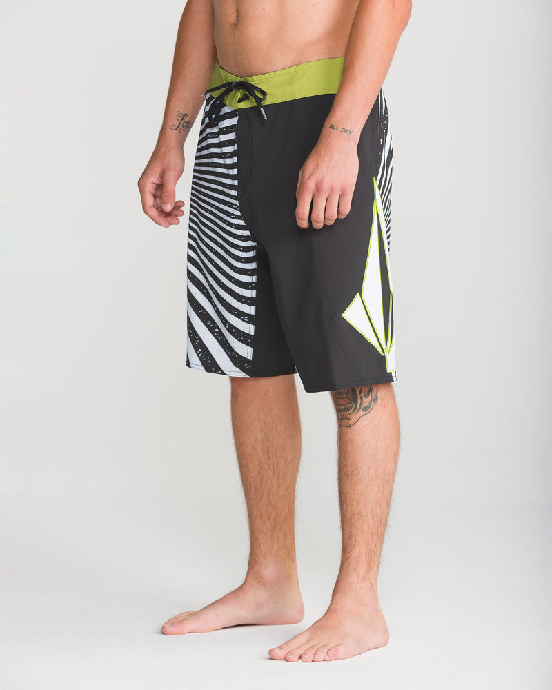 A man stands barefoot in Volcom’s Men’s Foster Mod 21 board shorts, featuring black and white stripes, a green waistband, and a yellow geometric design, crafted with 4-way stretch Mod-Tech fabric, against a plain background.