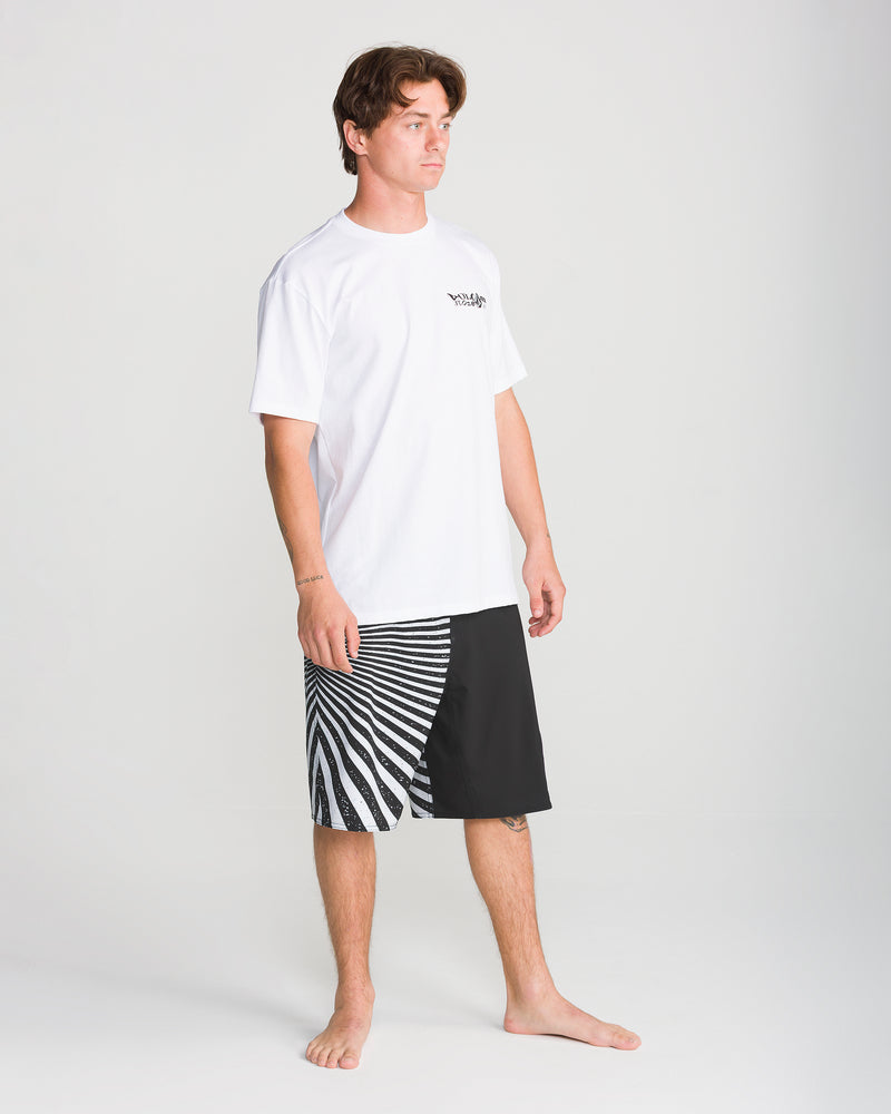 A person stands barefoot against a plain background, wearing a white T-shirt with a small logo and knee-length Volcom Men’s Foster Mod 21 shorts in black, which feature a distinctive white zebra-like pattern on one side. The shorts are designed with Mod-Tech construction and 4-way stretch for enhanced comfort and flexibility.