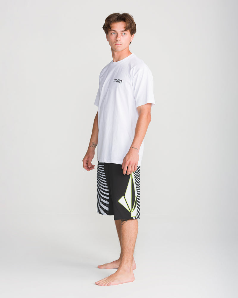 A young man with short brown hair is standing barefoot on a white background, wearing the Volcom Men’s Foster Mod 21 black shorts featuring Y2K styling, white stripes, and a yellow design, paired with a white T-shirt.
