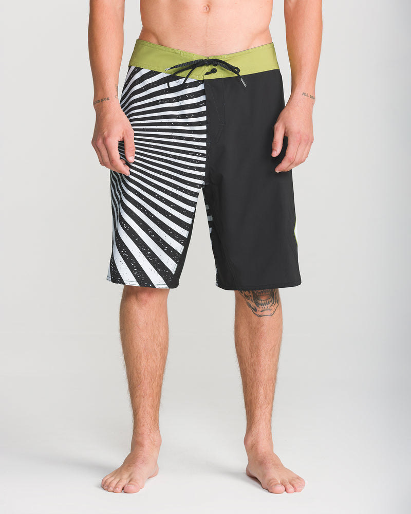 A person stands barefoot against a plain background, wearing Volcom Men’s Foster Mod 21 board shorts featuring a black and white pattern with a green waistband. Only the lower half of their body is visible.