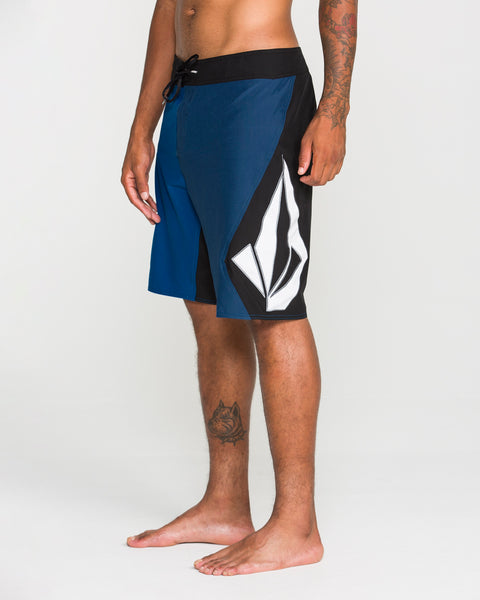 A person stands barefoot wearing the Volcom Men's Foster Mod 21" boardshorts in blue and black, featuring a large white geometric design on the side. The shorts have a loose fit and a convenient zip side pocket. Only the lower torso and legs are visible against a plain, light-colored background.