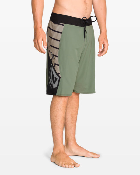 A person stands barefoot on a white background, wearing Volcom Men's Ripper Mod 21" green boardshorts with black and gray geometric patterns, a black drawstring waistband, and made from recycled polyester.