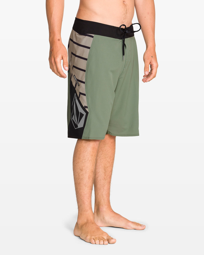 A person stands barefoot on a white background, wearing Volcom Men's Ripper Mod 21" green boardshorts with black and gray geometric patterns, a black drawstring waistband, and made from recycled polyester.