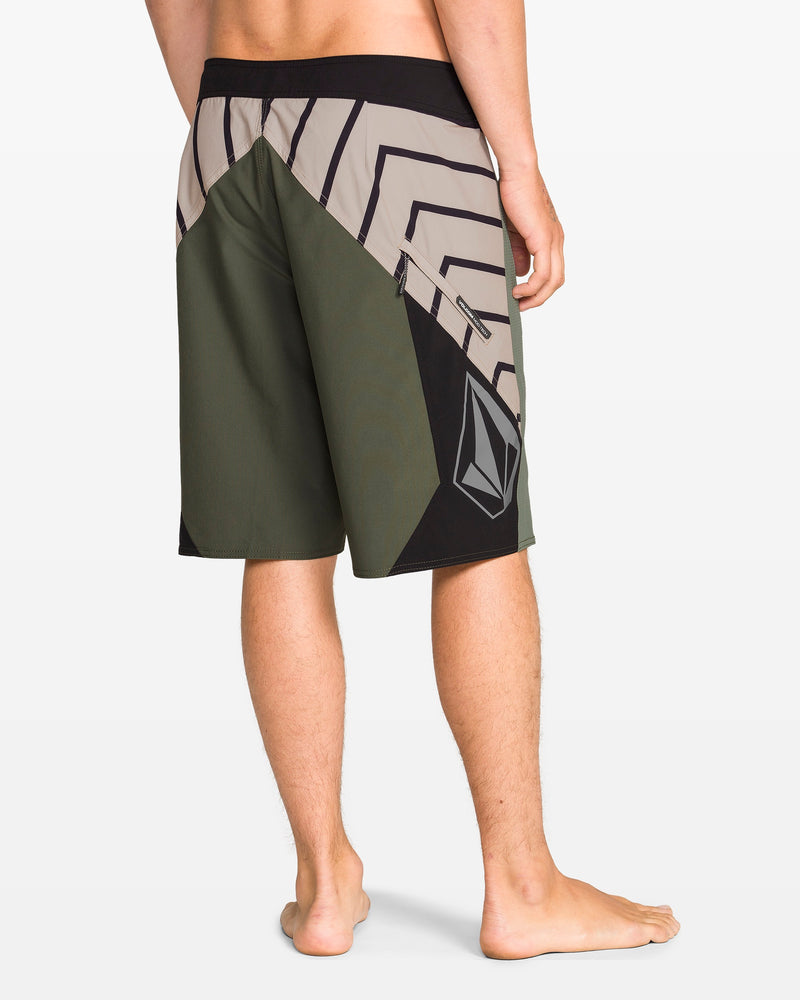 A person stands barefoot, facing away, wearing Volcom Men's Ripper Mod 21" boardshorts in green, black, and beige geometric patterns made from recycled polyester. The prominent Volcom logo is on the left leg against a plain white background.