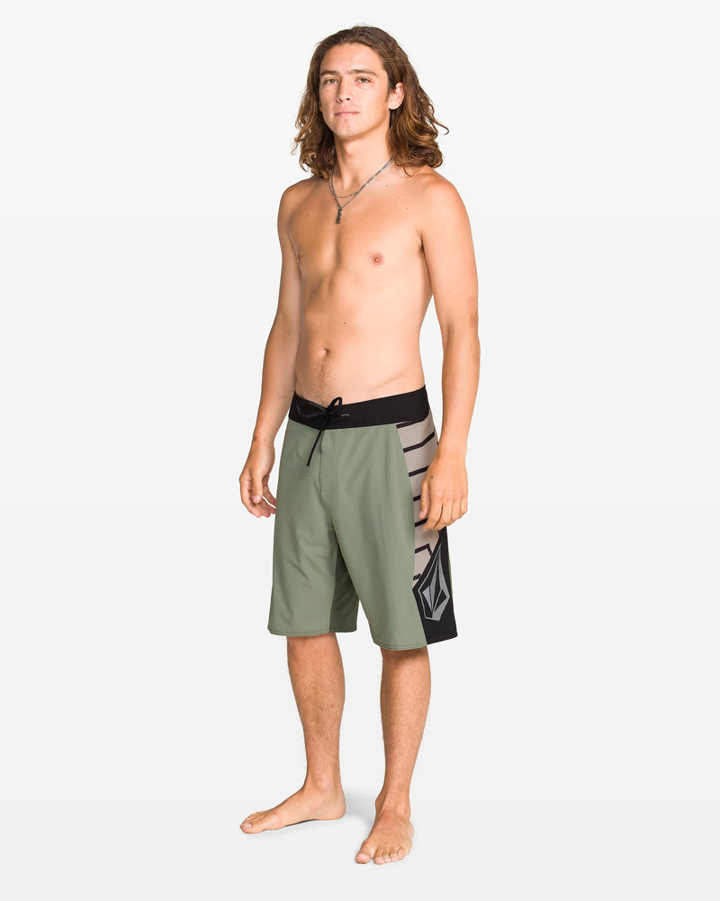 A young man with long brown hair stands barefoot against a white background, wearing Volcom Men's Ripper Mod 21" boardshorts made from recycled polyester. They are green with black and striped side panels. He is shirtless and looking slightly to his left.