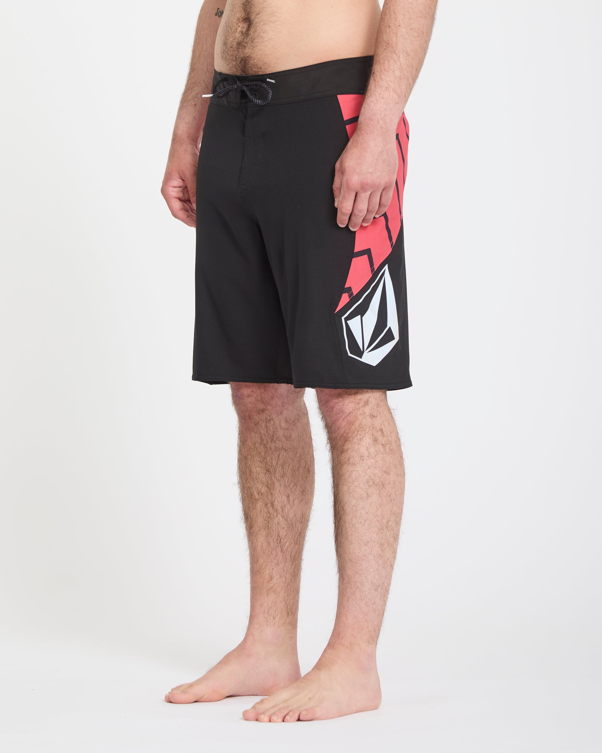 A person stands barefoot against a white background, wearing Volcom Men's Ripper Mod 21 black board shorts featuring a red graphic and white logo on the left side. The image is cropped from mid-torso to feet.