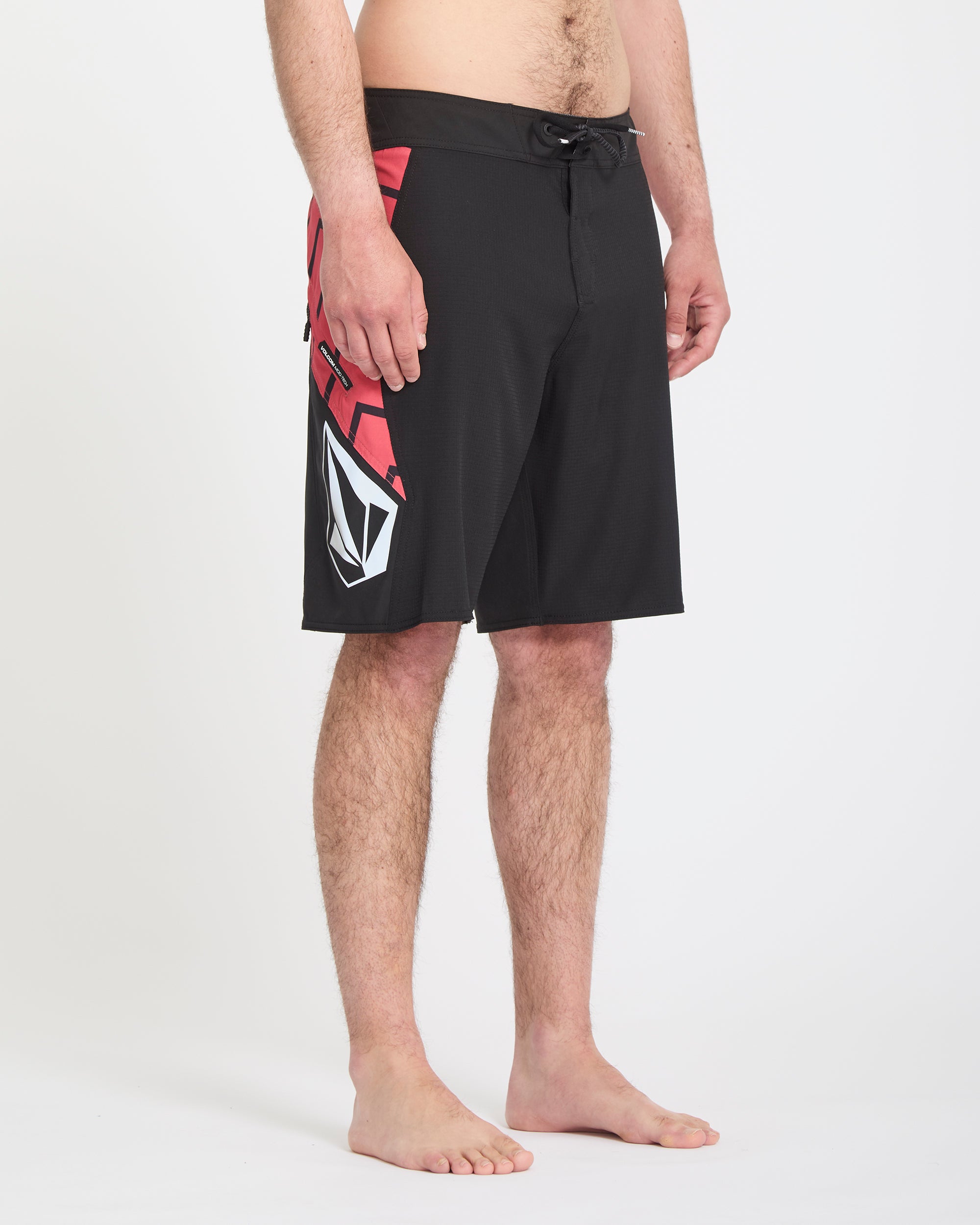 A person stands barefoot against a white background, wearing the Volcom Men's Ripper Mod 21 black board shorts featuring a red and white geometric pattern on the left side. The photo shows the model from waist to feet.
