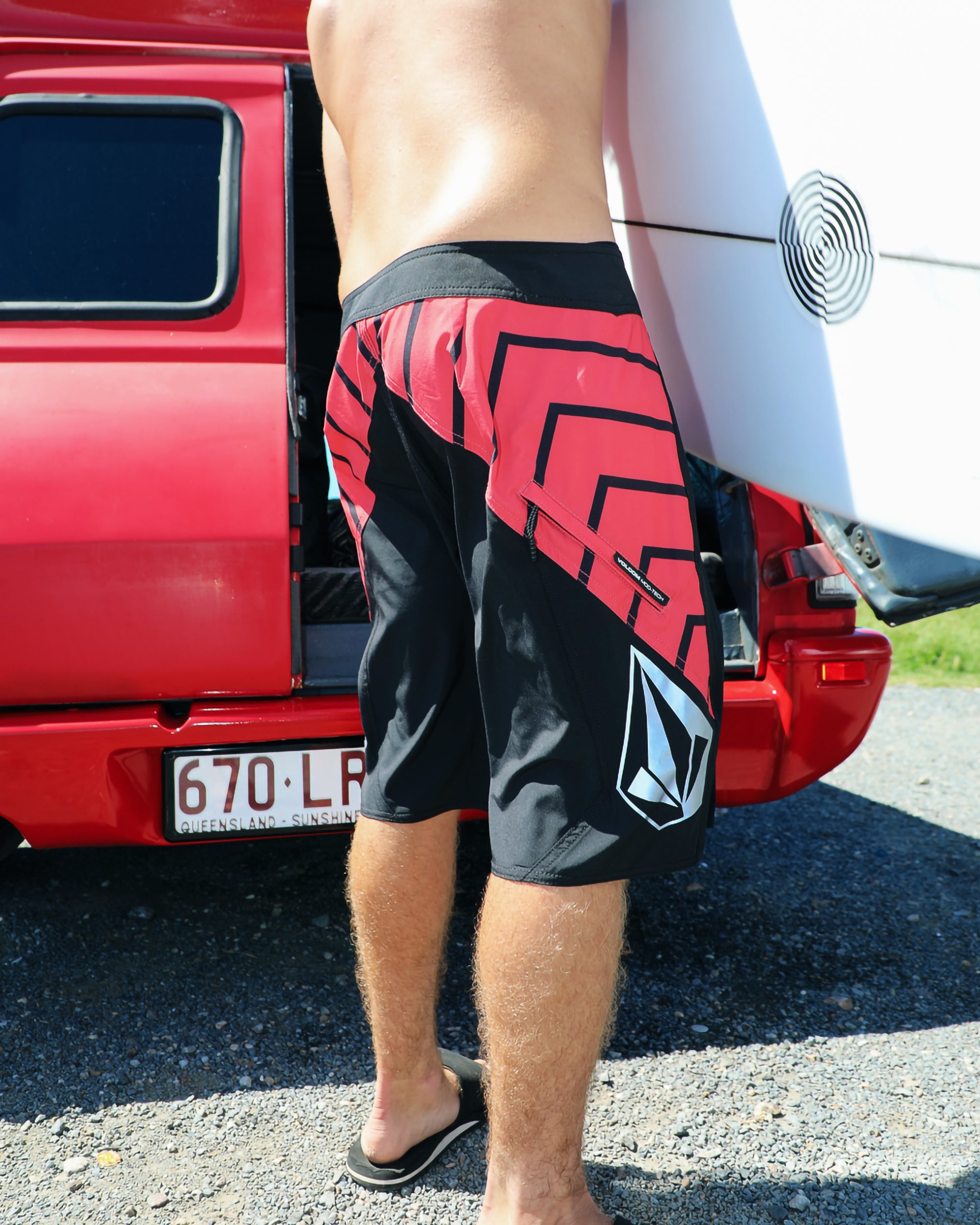 Wearing Volcom Men's Ripper Mod 21" black and red boardshorts made from recycled polyester, a person in flip-flops stands by an open-trunk red vehicle with a Queensland plate, holding a white surfboard.