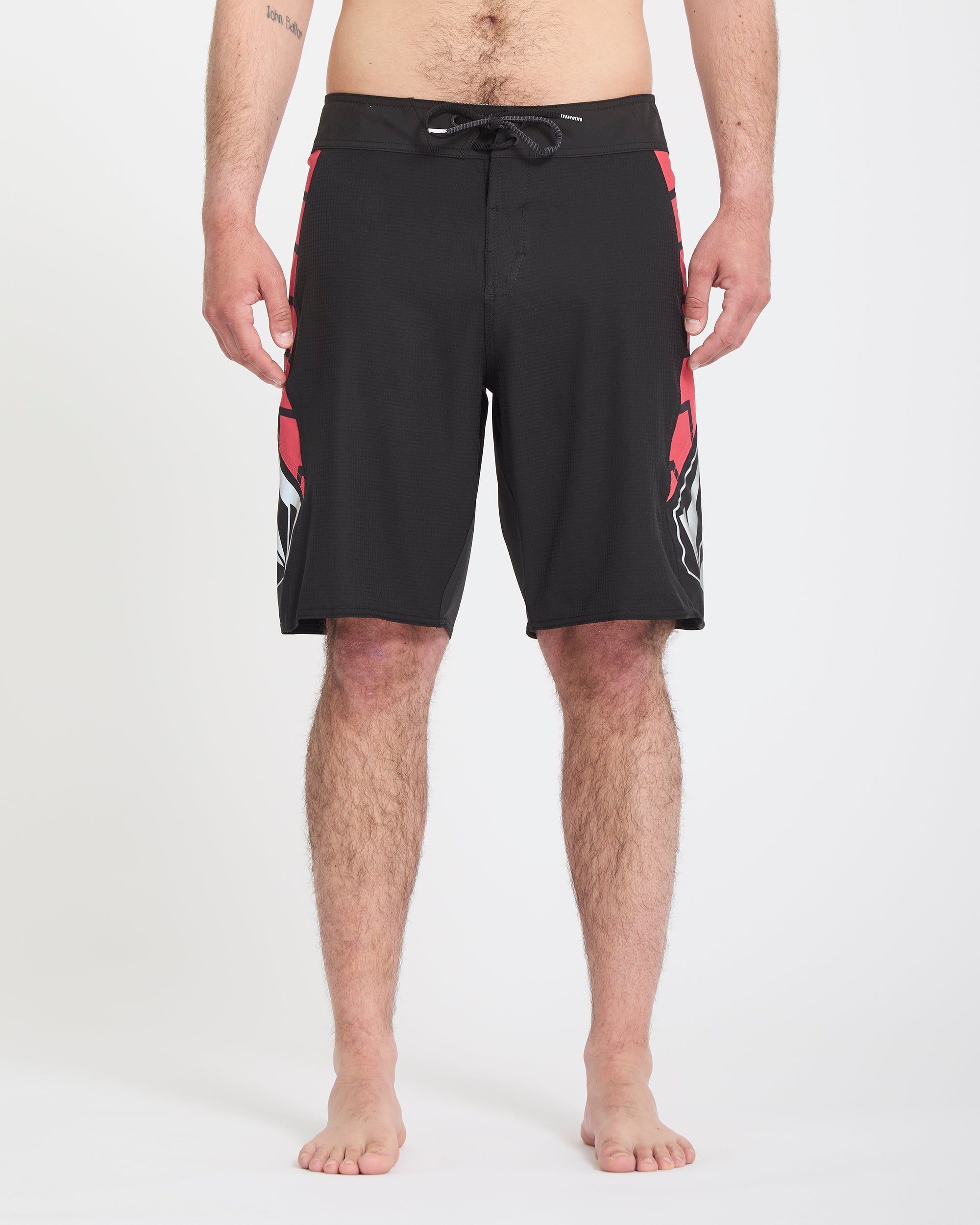 A barefoot man, shown from the chest down, models Volcom Men's Ripper Mod 21 board shorts featuring black fabric with red and white side designs, against a white background.