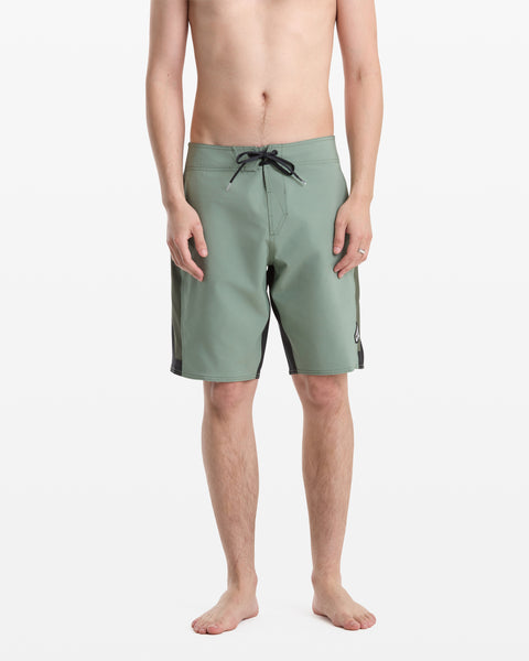 A person stands barefoot against a plain white background, wearing Volcom Men's React Mod 20 green knee-length board shorts with black drawstring and side panels. The person's upper body is uncovered.