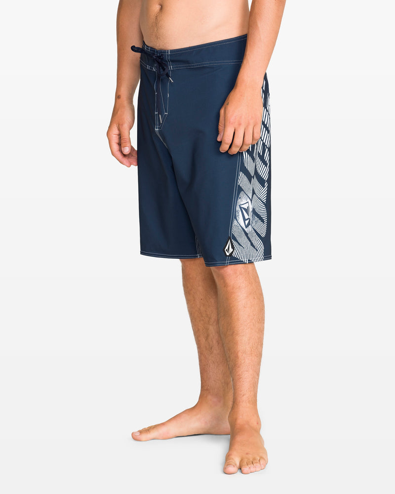 A man wears Volcom Men's Reaper Mod 21" boardshorts in midnight blue with a drawstring waist, geometric white side pattern, and REPREVE 4-way stretch fabric, standing barefoot against a plain white backdrop.