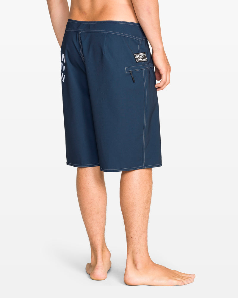 A barefoot person stands on a white background, shown from the waist down, wearing Volcom Men's Reaper Mod 21" boardshorts in midnight blue with a zippered pocket, REPREVE 4-way stretch fabric, and a small logo patch above the pocket.