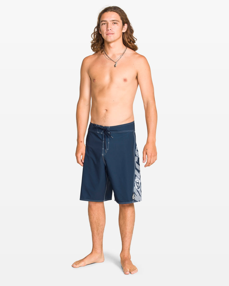 A young man with long hair stands barefoot against a white background, wearing Volcom Men's Reaper Mod 21" boardshorts in Midnight Blue with a graphic 90's design and REPREVE 4-way stretch. He is shirtless, wears a necklace, and looks at the camera.