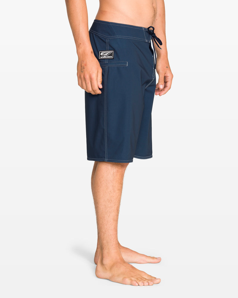 A person stands barefoot in profile, wearing Volcom Men's Reaper Mod 21" boardshorts in Midnight Blue with white stitching and a left pocket patch. The relaxed-fit shorts feature REPREVE 4-way stretch, a drawstring waist, and subtle 90's style on a white background.