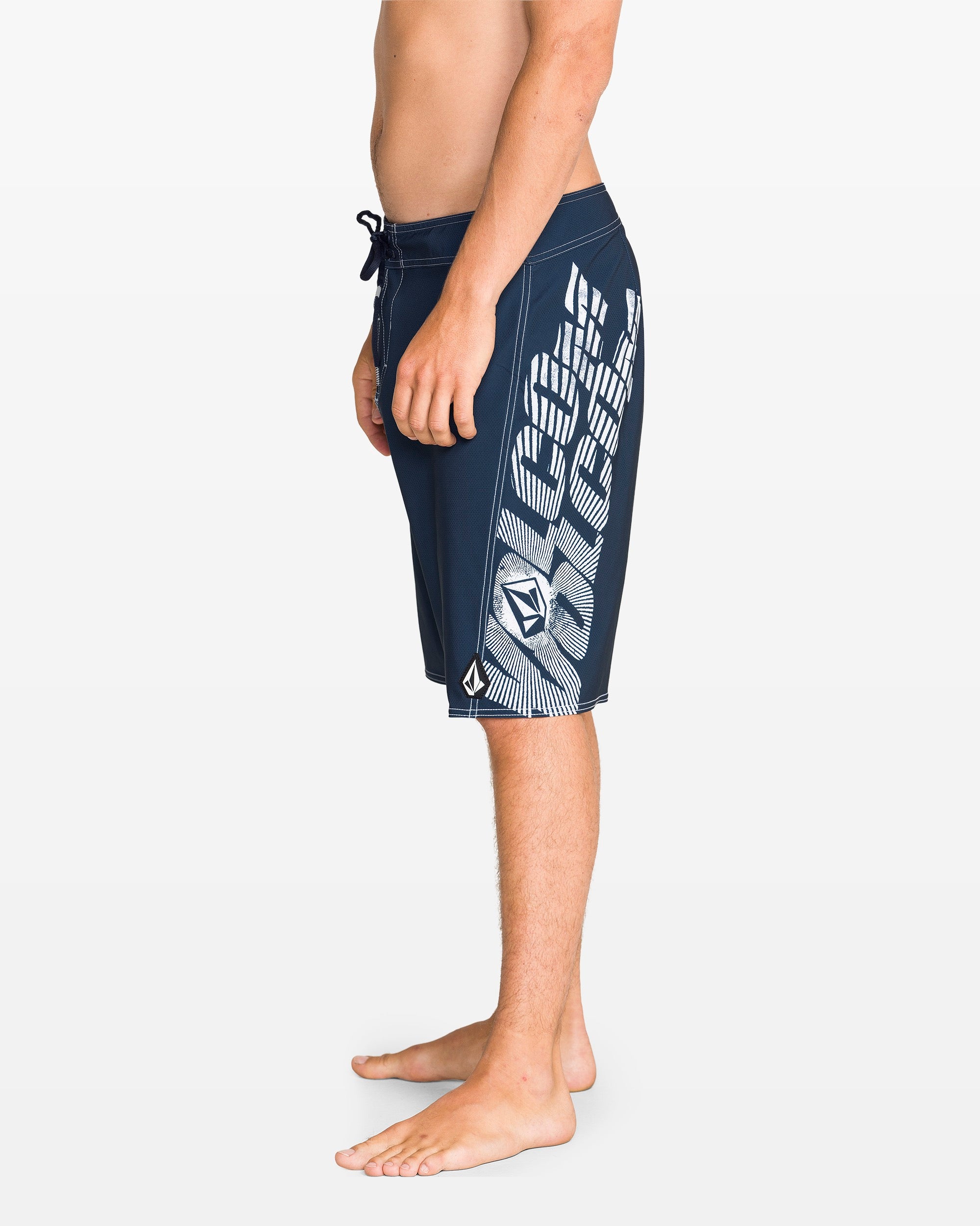 A man stands barefoot in profile, wearing Volcom Men's Reaper Mod 21" boardshorts in Midnight Blue with 90's style white geometric patterns and a drawstring waist, made from REPREVE 4-way stretch fabric. The background is plain white.