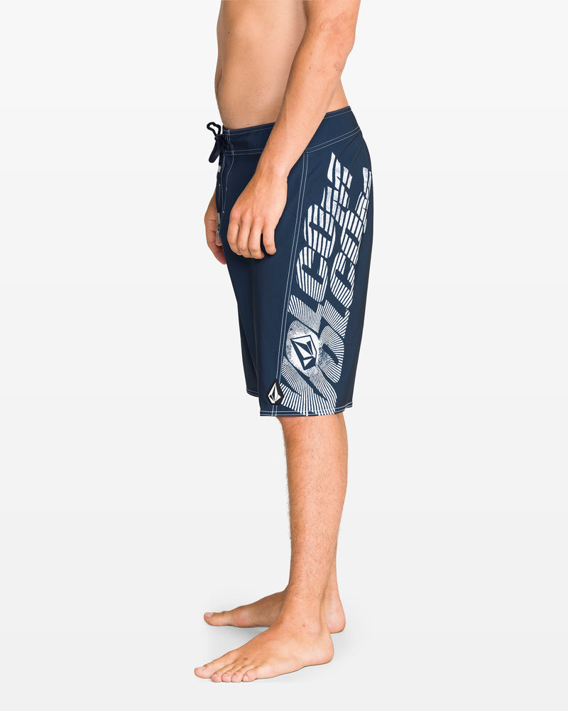 A man stands barefoot in profile, wearing Volcom Men's Reaper Mod 21" boardshorts in Midnight Blue with 90's style white geometric patterns and a drawstring waist, made from REPREVE 4-way stretch fabric. The background is plain white.