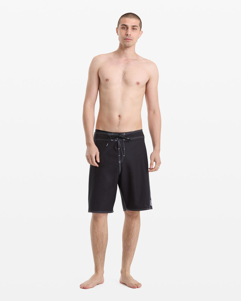 A young man with short hair stands barefoot against a white background, wearing Volcom Men's Reaper Mod 21 black board shorts with a drawstring. He faces forward with a neutral expression and relaxed posture.