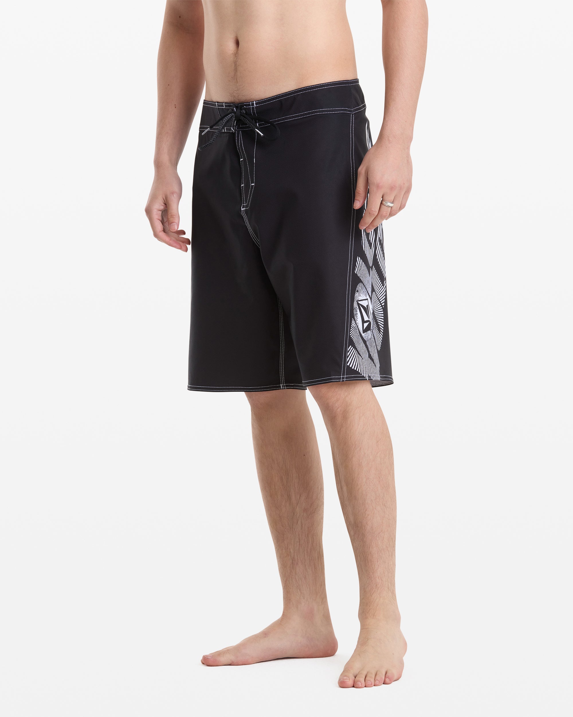 A person models Volcom Men's Reaper Mod 21 black board shorts with white stitching and a geometric side pattern, standing barefoot against a white background with face not shown.
