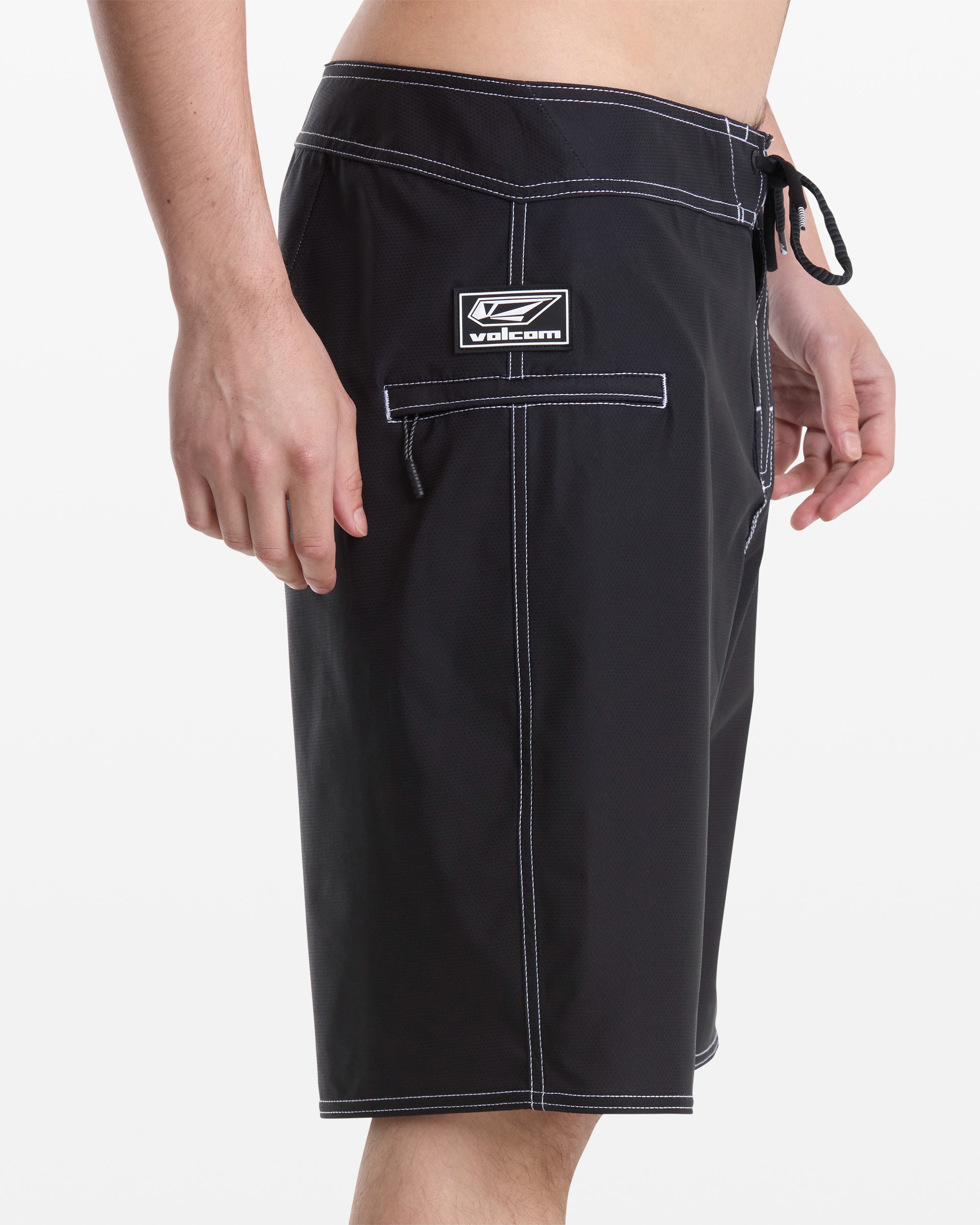 A man stands sideways to the camera wearing Volcom Men's Reaper Mod 21 black board shorts with white stitching, featuring a side pocket with a Velcro flap and a logo patch above it.