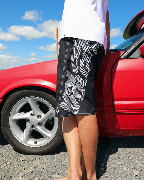 A person wearing Volcom Men's Reaper Mod 21" black boardshorts made from recycled polyester and a white shirt stands barefoot beside a red car on gravel under a blue sky with scattered clouds.