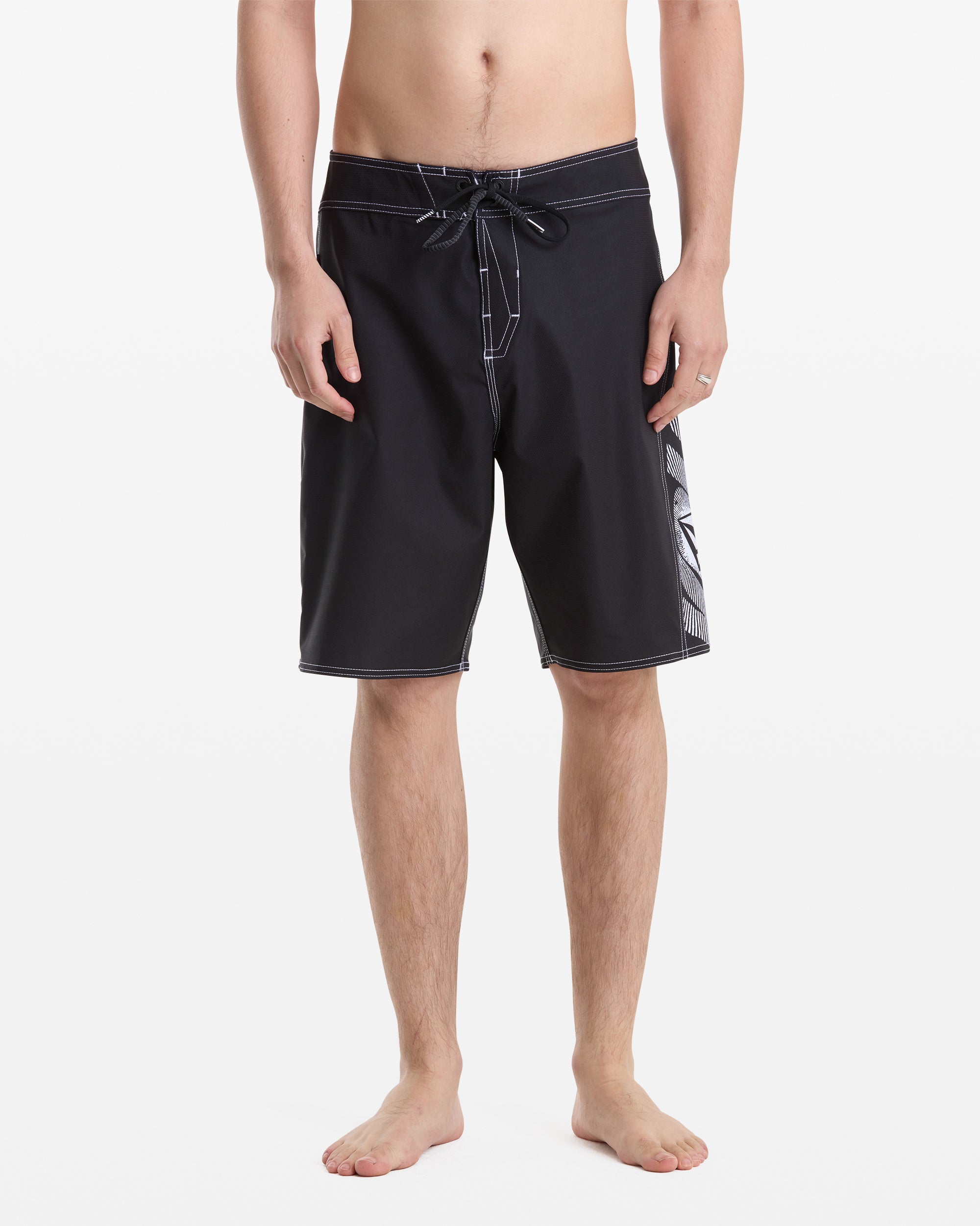 A barefoot man models Volcom’s Men's Reaper Mod 21 black board shorts with white stitching, drawstring, and a patterned side panel. Photo shows him from the waist down against a white background.
