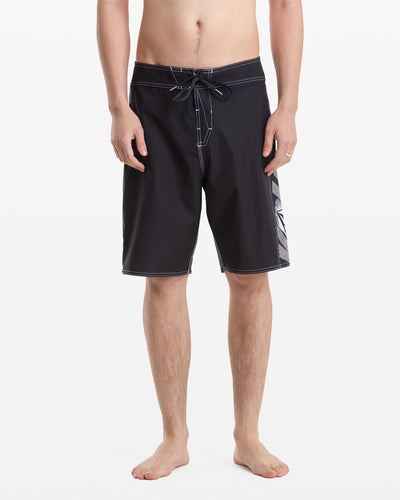 A barefoot man models Volcom’s Men's Reaper Mod 21 black board shorts with white stitching, drawstring, and a patterned side panel. Photo shows him from the waist down against a white background.