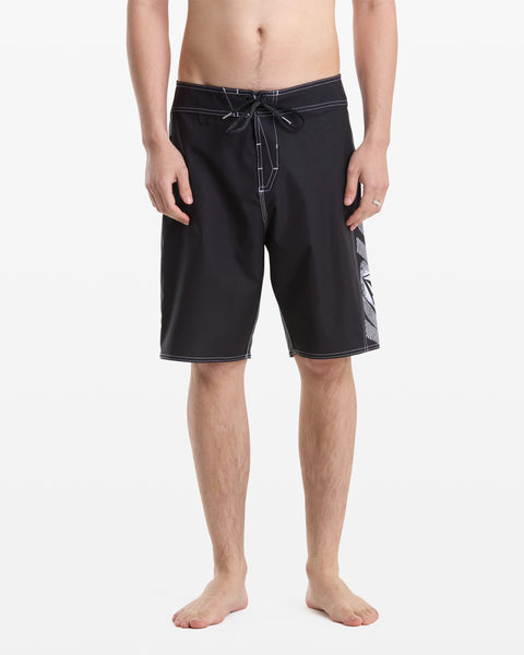 A barefoot man models Volcom’s Men's Reaper Mod 21 black board shorts with white stitching, drawstring, and a patterned side panel. Photo shows him from the waist down against a white background.