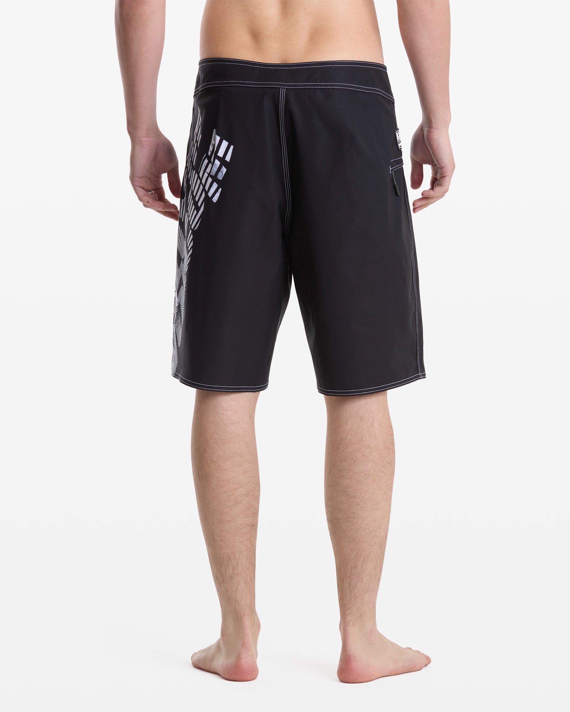 A man stands barefoot, facing away from the camera, wearing Volcom Men's Reaper Mod 21 knee-length black shorts with white stitching and a geometric white pattern on the left side against a plain white background.