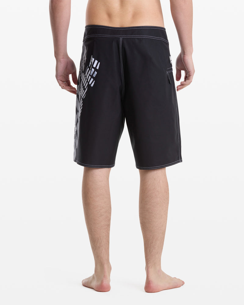 A man stands barefoot, facing away from the camera, wearing Volcom Men's Reaper Mod 21 knee-length black shorts with white stitching and a geometric white pattern on the left side against a plain white background.
