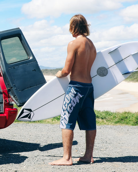 A barefoot man in Volcom’s Men’s Reaper Mod Midnight Blue boardshorts made with REPREVE 4-way stretch stands by a red vehicle, holding a white surfboard and facing the beach on a sunny day.