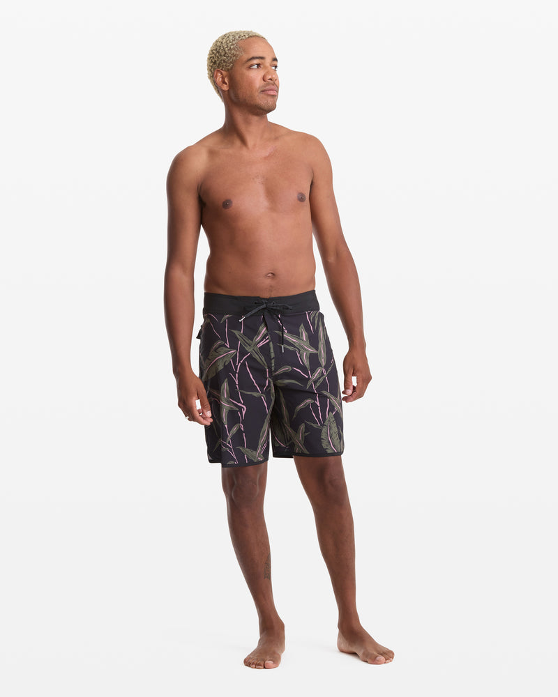 A barefoot man with short, bleached blond hair stands against a white background, wearing Volcom Men's Encounter Scallop Mod 19 black shorts with a colorful leaf pattern. He is shirtless and looking slightly to his left.