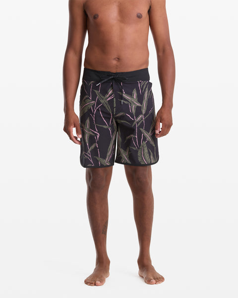 A person stands barefoot against a white background, wearing Volcom Men's Encounter Scallop Mod 19 black swim trunks with green and purple tropical leaf patterns. The image is cropped from the chest down.