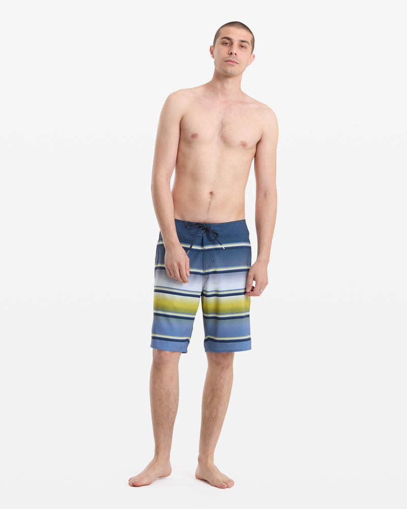 A young man stands barefoot against a white background, wearing Volcom Men's Quarta Dissolve Mod 20 board shorts with blue, green, and white stripes. He is shirtless with a relaxed posture and neutral expression.