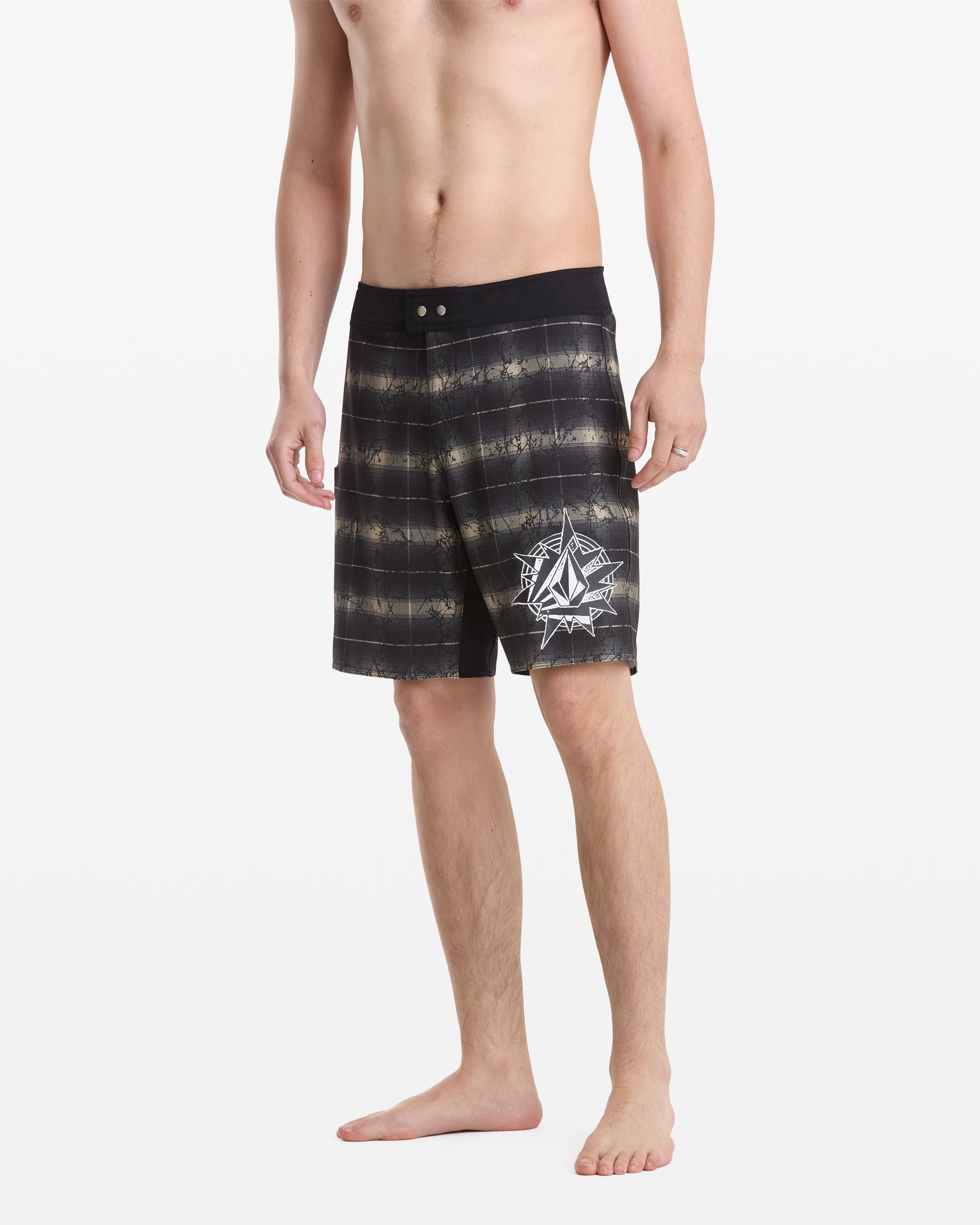 A man stands barefoot in Volcom Men's Grunge Snap Mod 18 board shorts, featuring black and gray stripes, a white geometric compass on the left leg, a black waistband, and a knee-length cut.