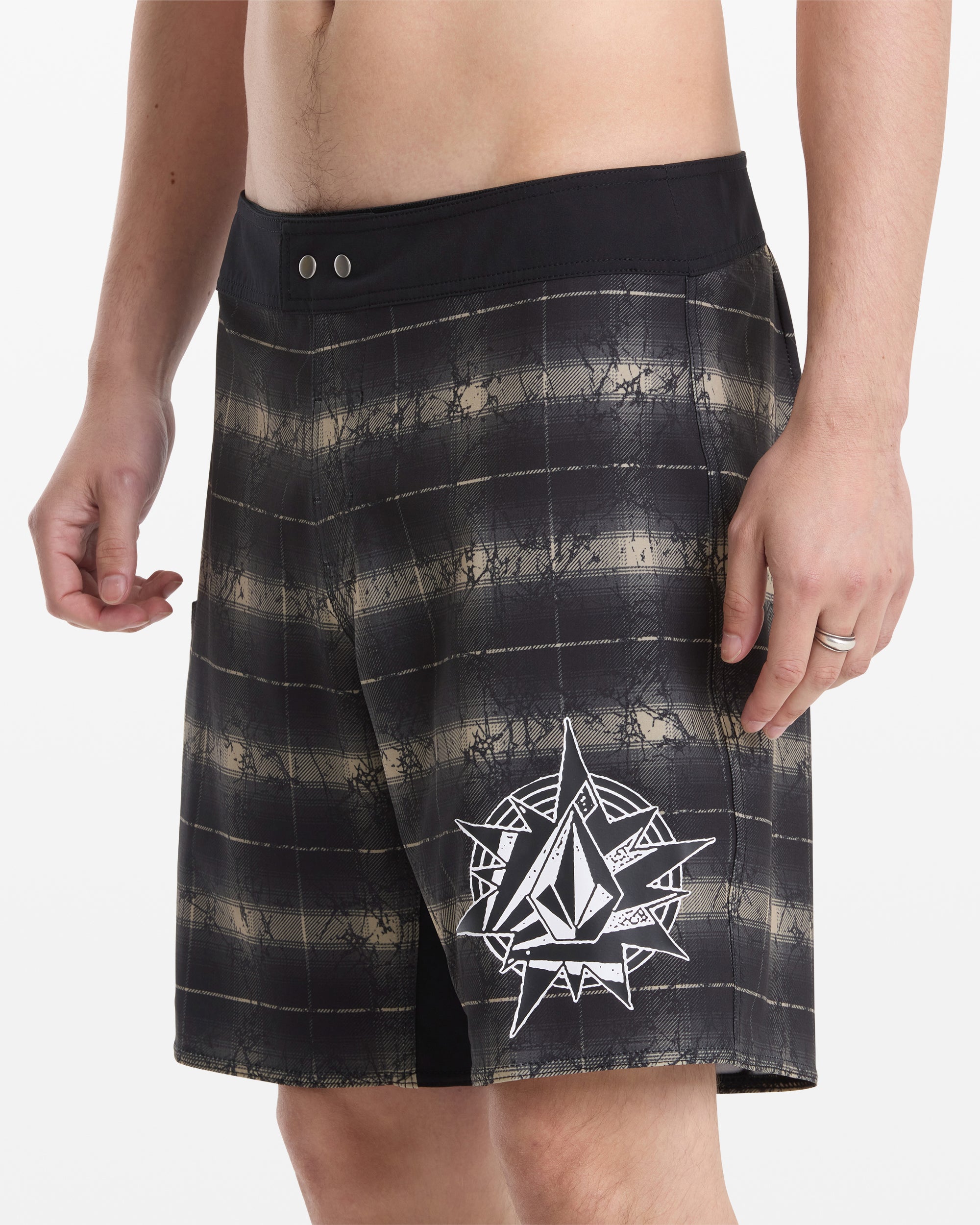 A man wears Volcom Men's Grunge Snap Mod 18 black and grey plaid board shorts with a large white geometric logo on the left leg, standing against a plain white background.