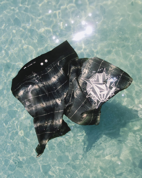 A pair of Volcom Men's Grunge Snap Mod 18 black plaid swim trunks with a white geometric design float on clear blue water under bright sunlight.