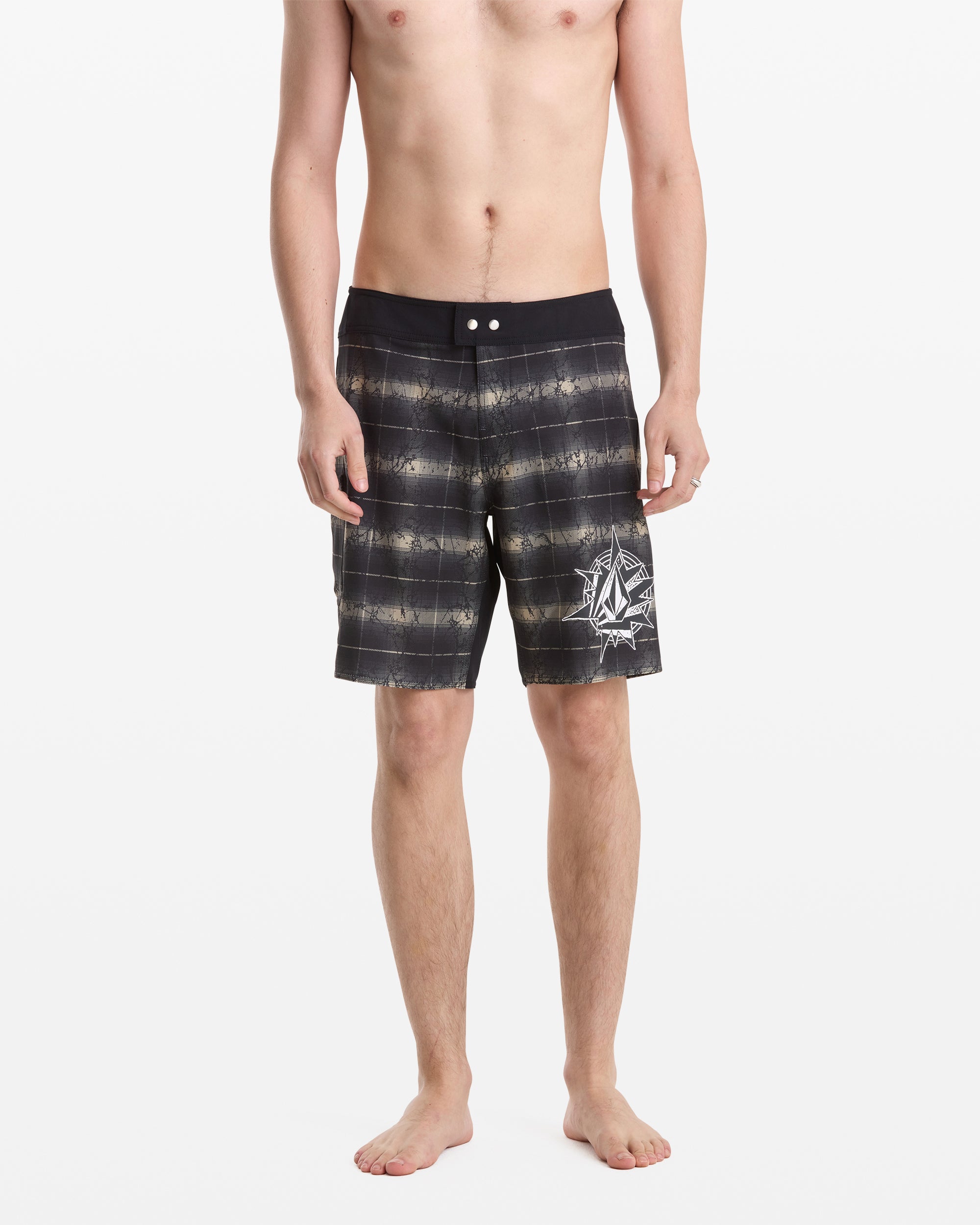 A shirtless man stands barefoot against a white background wearing Volcom Men’s Grunge Snap Mod 18 board shorts, featuring black and gray stripes and a white abstract graphic on the left leg.