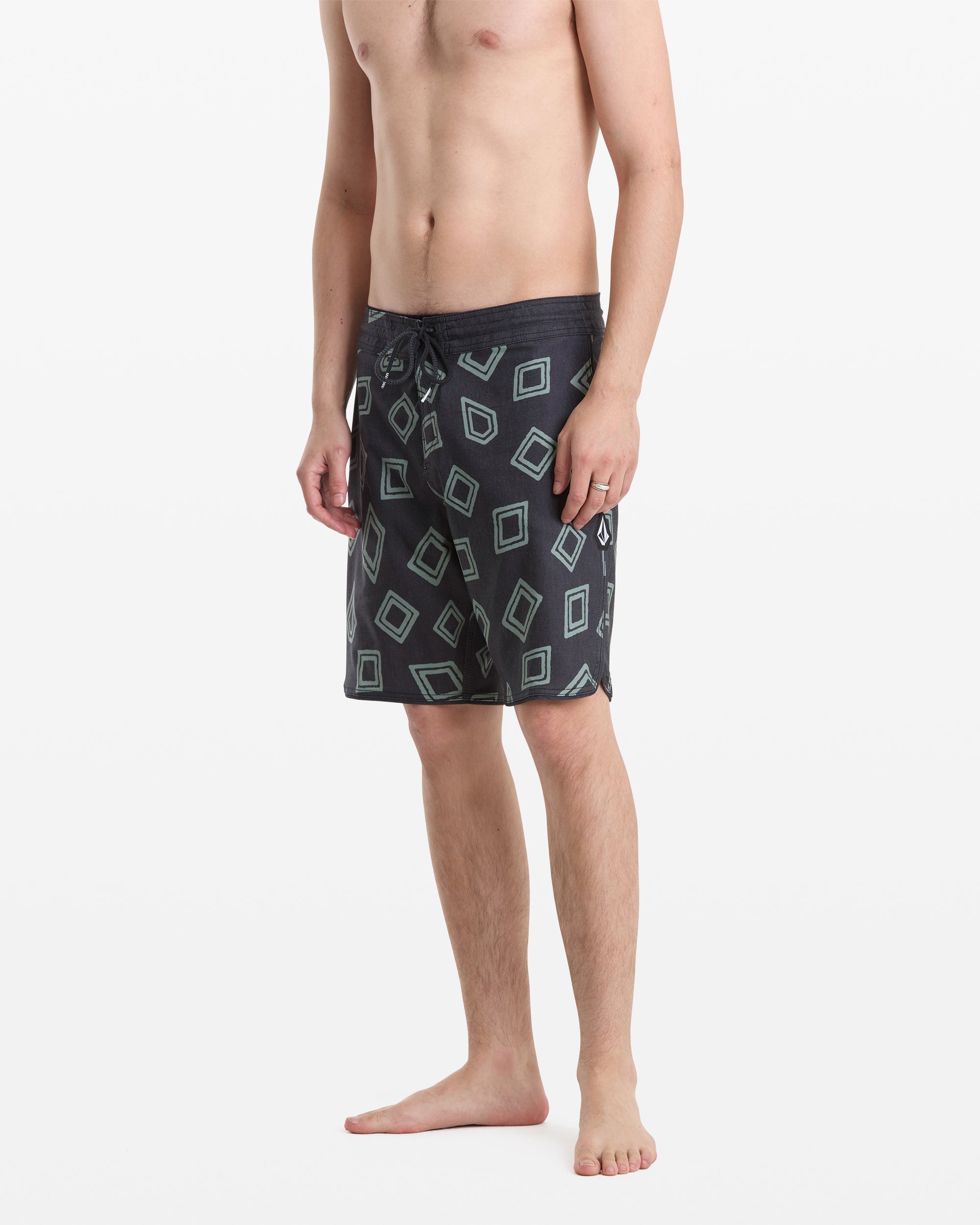 A man stands barefoot, shirtless, wearing Volcom Men's Contact Scallop Stoney 19 swim trunks with a geometric square pattern, facing slightly to the side against a plain white background.
