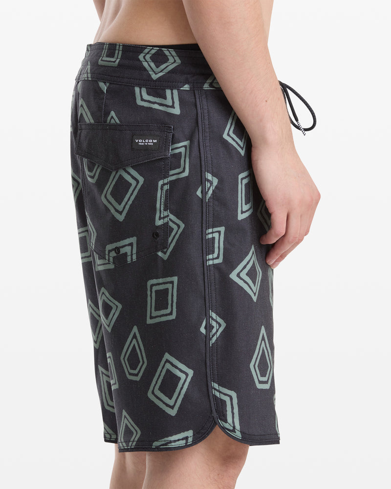 A man wearing Volcom Men's Contact Scallop Stoney 19 black shorts with a green geometric pattern and a back pocket featuring the Volcom logo patch, shown from the side against a plain white background.