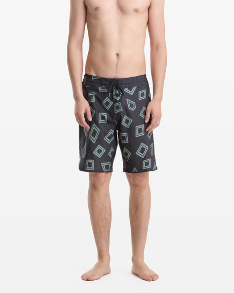 A man stands barefoot wearing Volcom Men's Contact Scallop Stoney 19 black swim trunks with a green square geometric pattern, shown from the waist down against a plain white background.