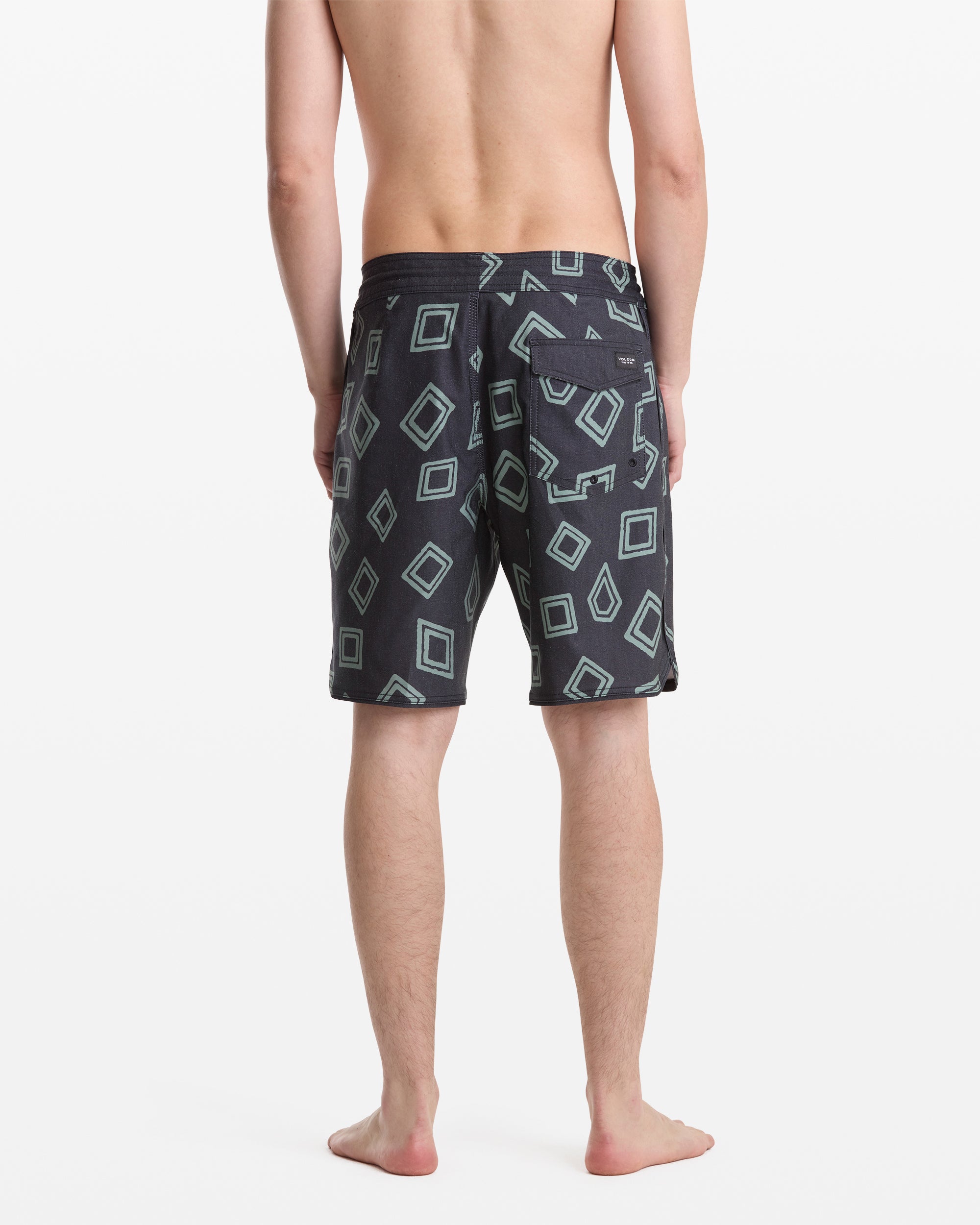 A man stands barefoot facing away, wearing Volcom Men's Contact Scallop Stoney 19 swim trunks in dark color with light blue geometric square patterns on a white background.