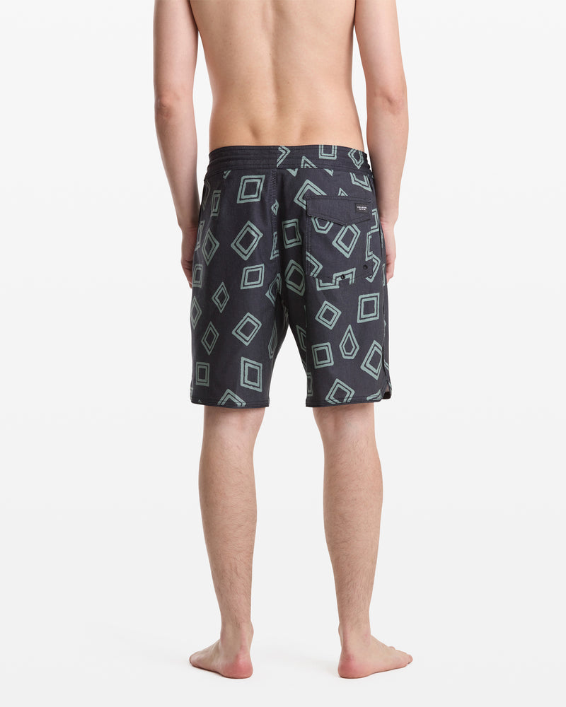 A man stands barefoot facing away, wearing Volcom Men's Contact Scallop Stoney 19 swim trunks in dark color with light blue geometric square patterns on a white background.