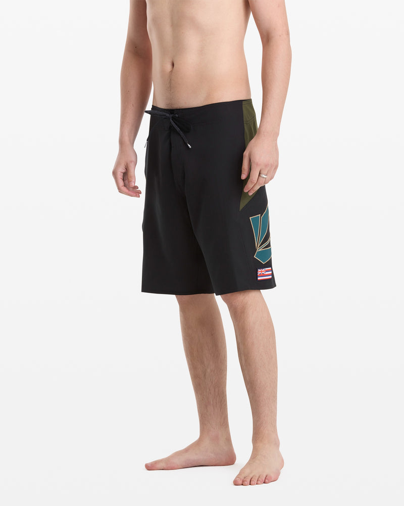 A man stands barefoot in Volcom Men's Hawaii Top Tier Mod black board shorts with a drawstring waist and a small graphic on the left leg; his torso is partially out of frame.
