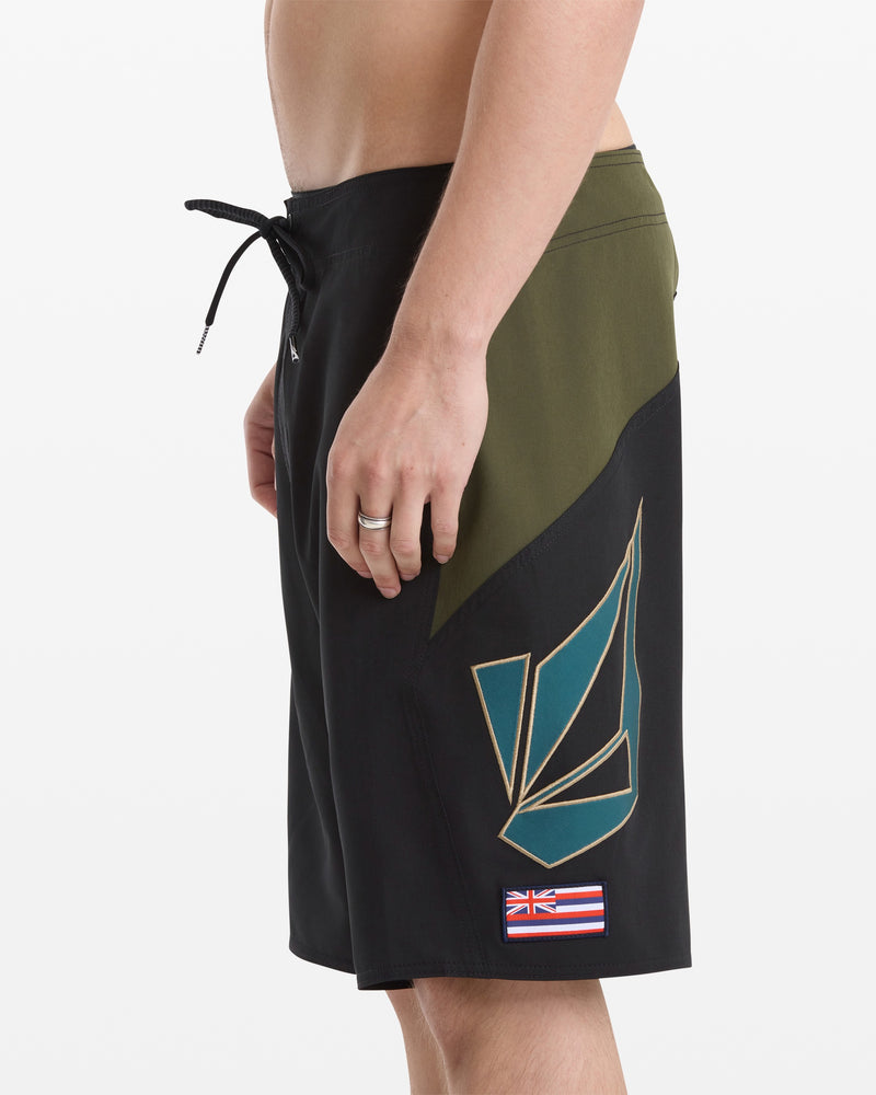 A man wears Volcom Men's Hawaii Top Tier Mod board shorts in black and olive green with a geometric pattern and Hawaiian flag patch on the left leg, standing against a plain white background.