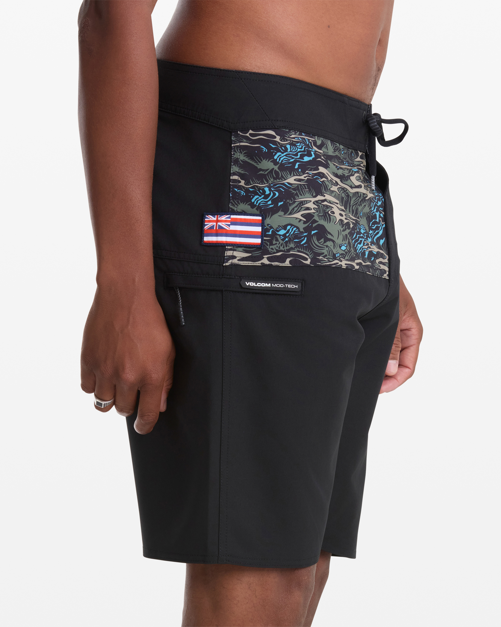 A person wearing Volcom Men's Parillo Mod 21 boardshorts, featuring black fabric and a camouflage-patterned pocket with a small Hawaiian flag patch, plus a black drawstring waist. Their arm rests relaxed at their side.