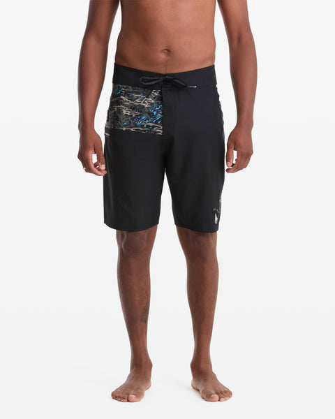 A person stands barefoot wearing Volcom Men's Parillo Mod 21 black board shorts featuring a colorful abstract panel on the upper front, against a plain white background.