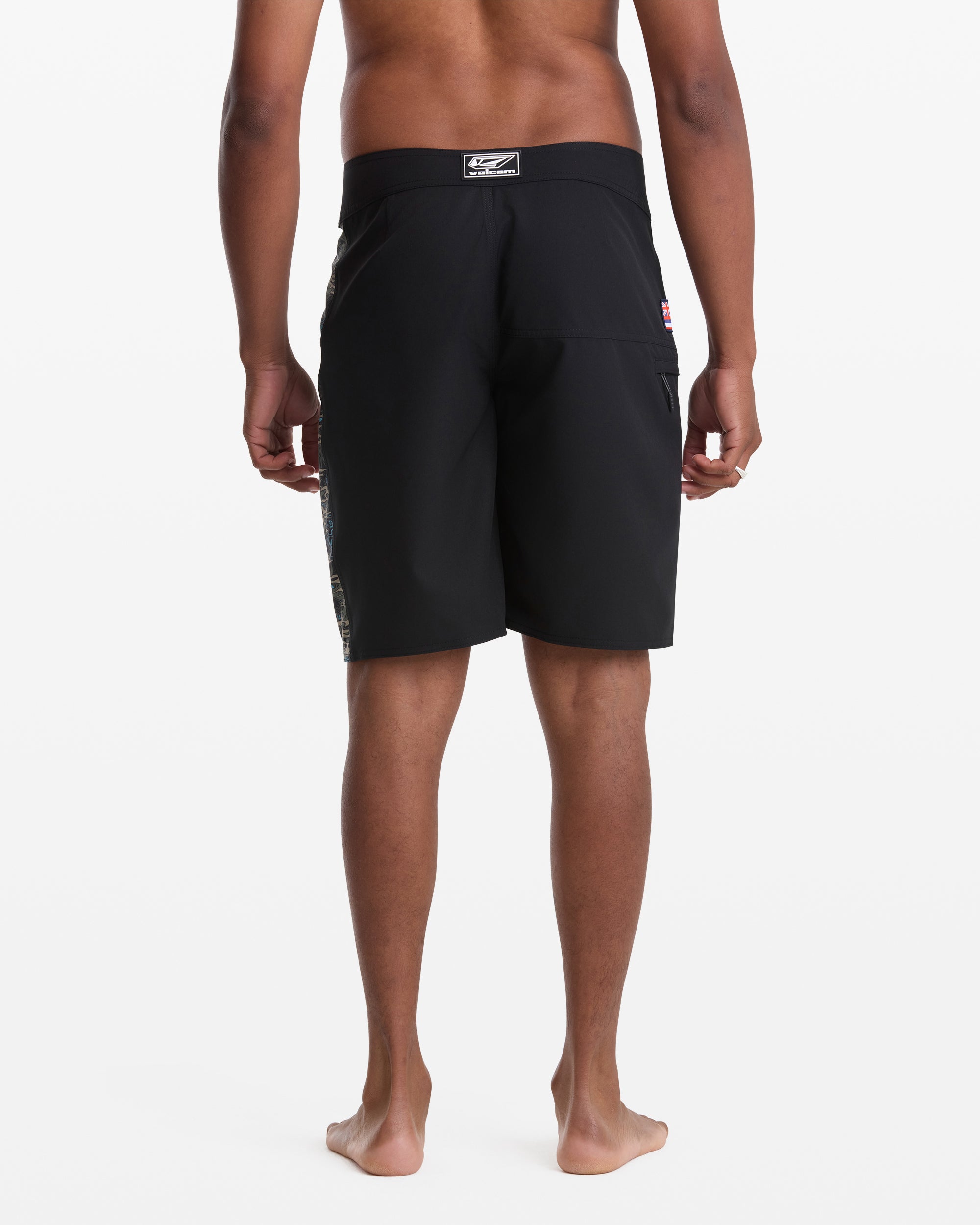A man stands barefoot facing away from the camera, wearing Volcom Men's Parillo Mod 21 black board shorts with a patch on the waistband and a side pocket. The background is white.