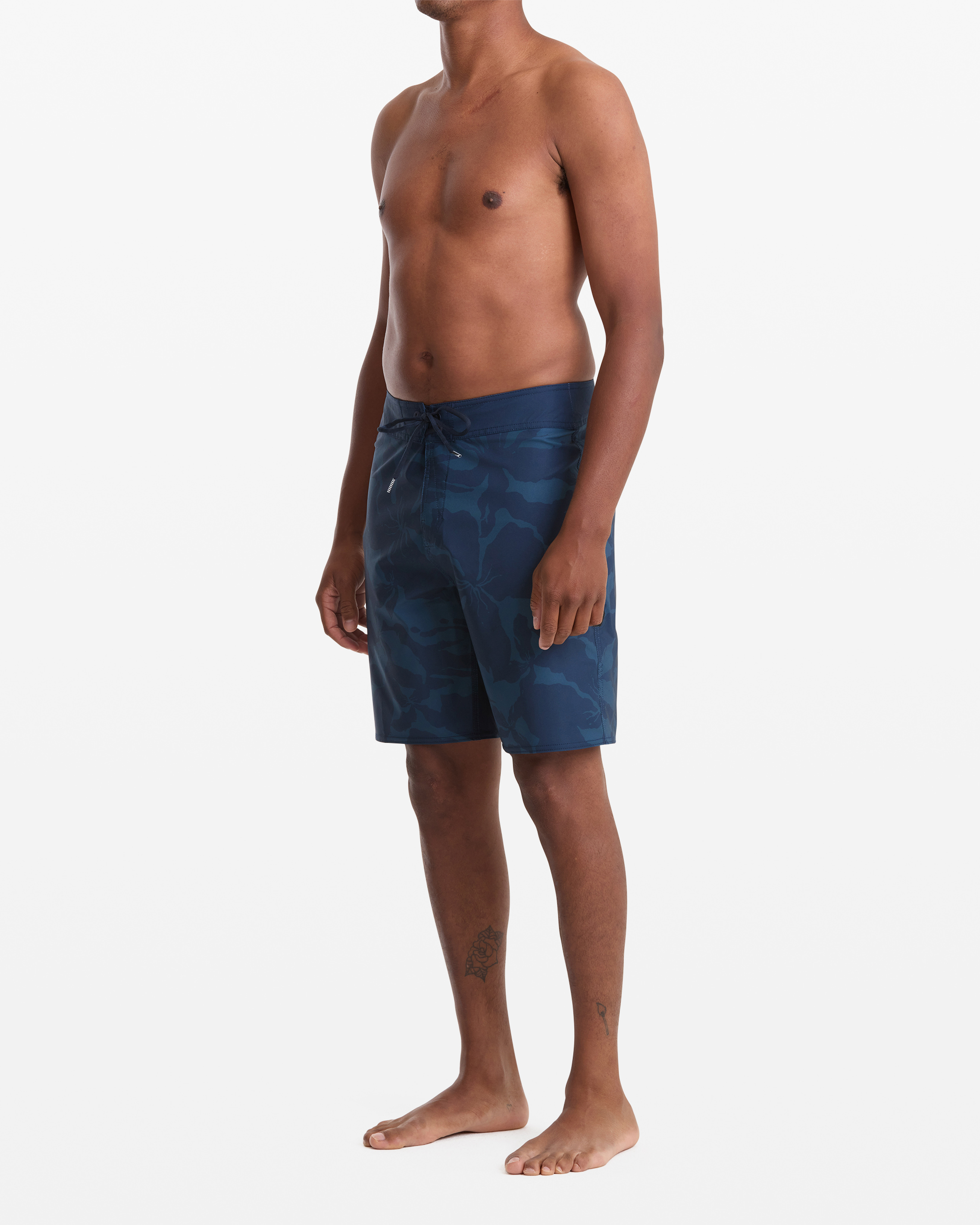 A man stands barefoot against a white background, wearing Volcom Men's Shoreline Mod 20 blue camouflage board shorts. He's shirtless, facing slightly left, with his arms relaxed at his sides.