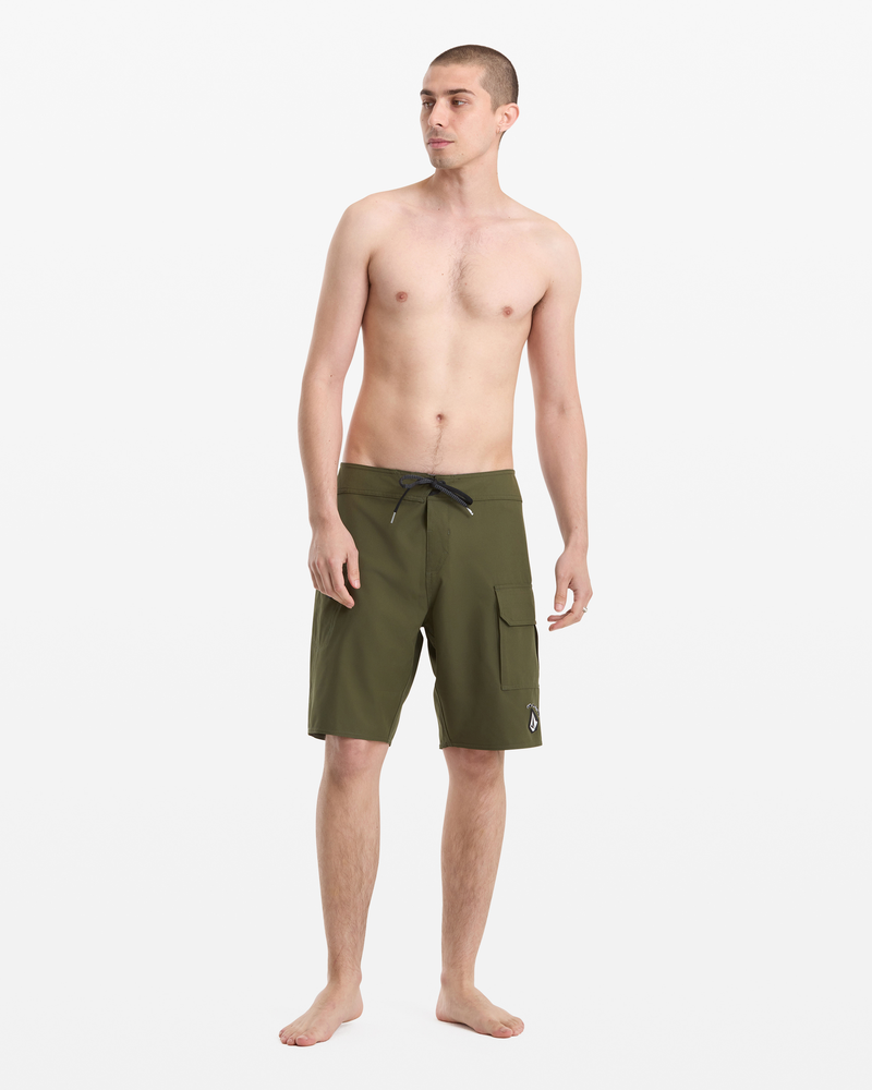 A barefoot man with short hair faces slightly right, modeling Volcom Men's Hawaii Truly Preemo Mod 20 olive green board shorts with a black drawstring and side pocket, on a plain white background.