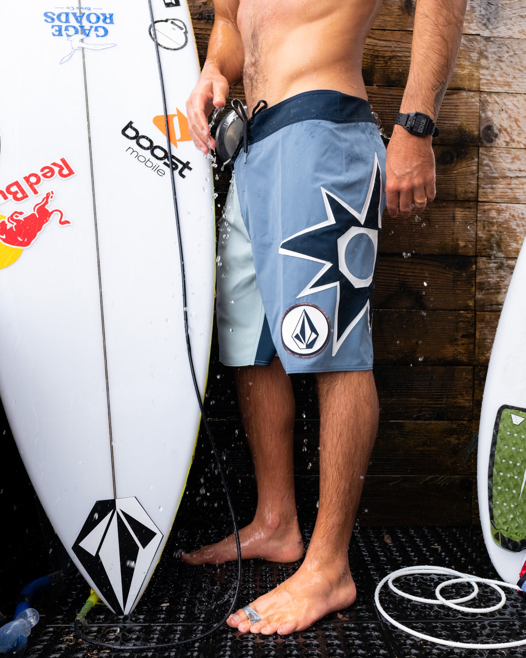 A man wears Volcom Men's St4R G4Zer Mod blue and gray boardshorts, standing by a stickered surfboard under running water near a wooden wall, with another surfboard partially visible on the right.